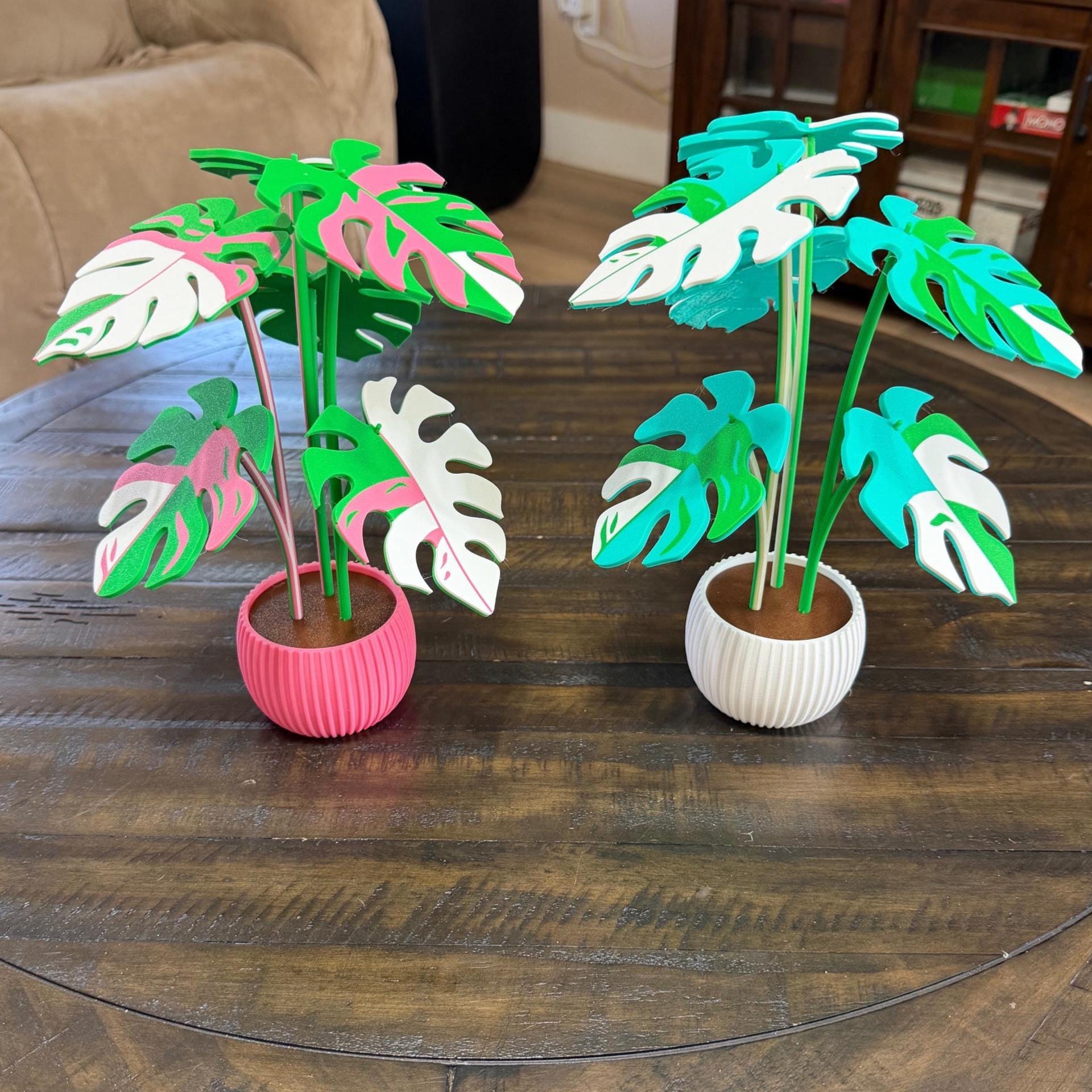 May include: Two decorative artificial Monstera plants. One plant has pink and white leaves in a pink pot, the other has blue and white leaves in a white pot. Both plants have green stems and are on a wooden table.